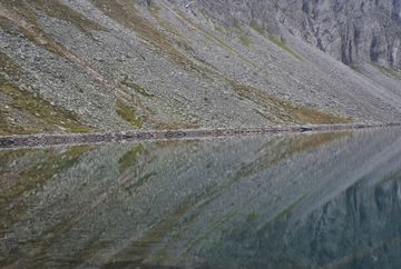 Das felsige Ufer spiegelt sich in der glatten Wasserfläche eines Bergsees