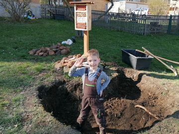 Ein kleiner Junge steht in einem Loch auf der Wiese, welches er ausgegraben hat