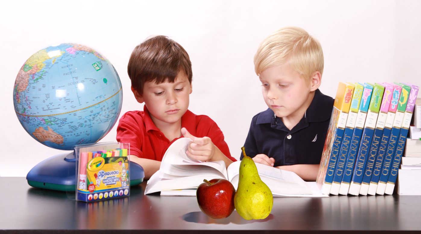 Zwei Buben an einem Tisch mit Globus und Büchern vergleichen einen Apfel mit einer Birne 